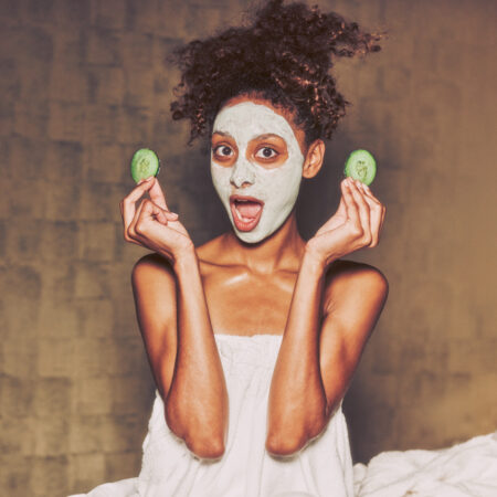 Seated woman with a facial mask and wrapped in a towel cheekily holds cucumber slices in each hand.