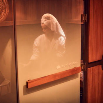 Woman relaxing in an infrared sauna in a robe with a wrap on her head, as viewed through the sauna door.