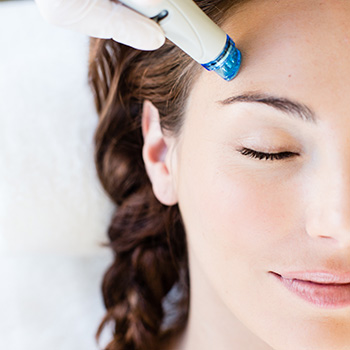 A woman lying on her back with her closed eyes with the Hydrafacial cleansing tip on her forehead.