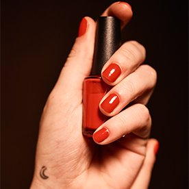 Woman's hand wrapped around a red bottle of nail polish, with red manicure and small moon tattoo