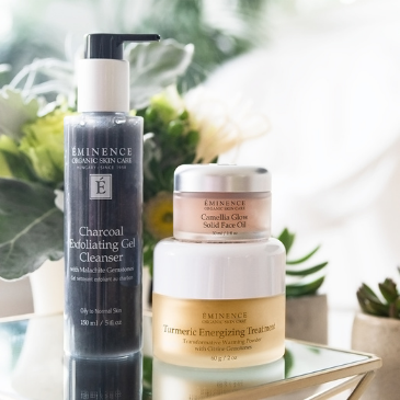 Three organic skin care products from Eminence Organics on a table, including Charcoal Exfoliating Gel Cleanser, Camilla Glow Solid Face Oil, and Tumeric Energizing Treatment.