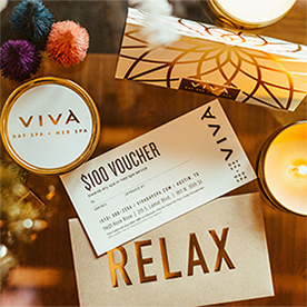Gold-foiled Viva Day Spa gift certificate with a Bonus $100 Holiday Voucher laying on a table next to an envelope with the words 